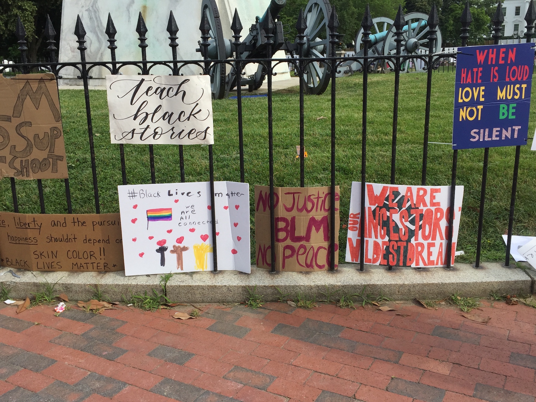 Hand-drawn protest signs hang on black bars of a fence in front of a green lawn. 