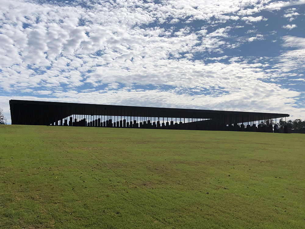 Outside of the National Memorial for Peace and Justice, Montgomery, Alabama, with a grassy field in front, blue sky with scattered clouds