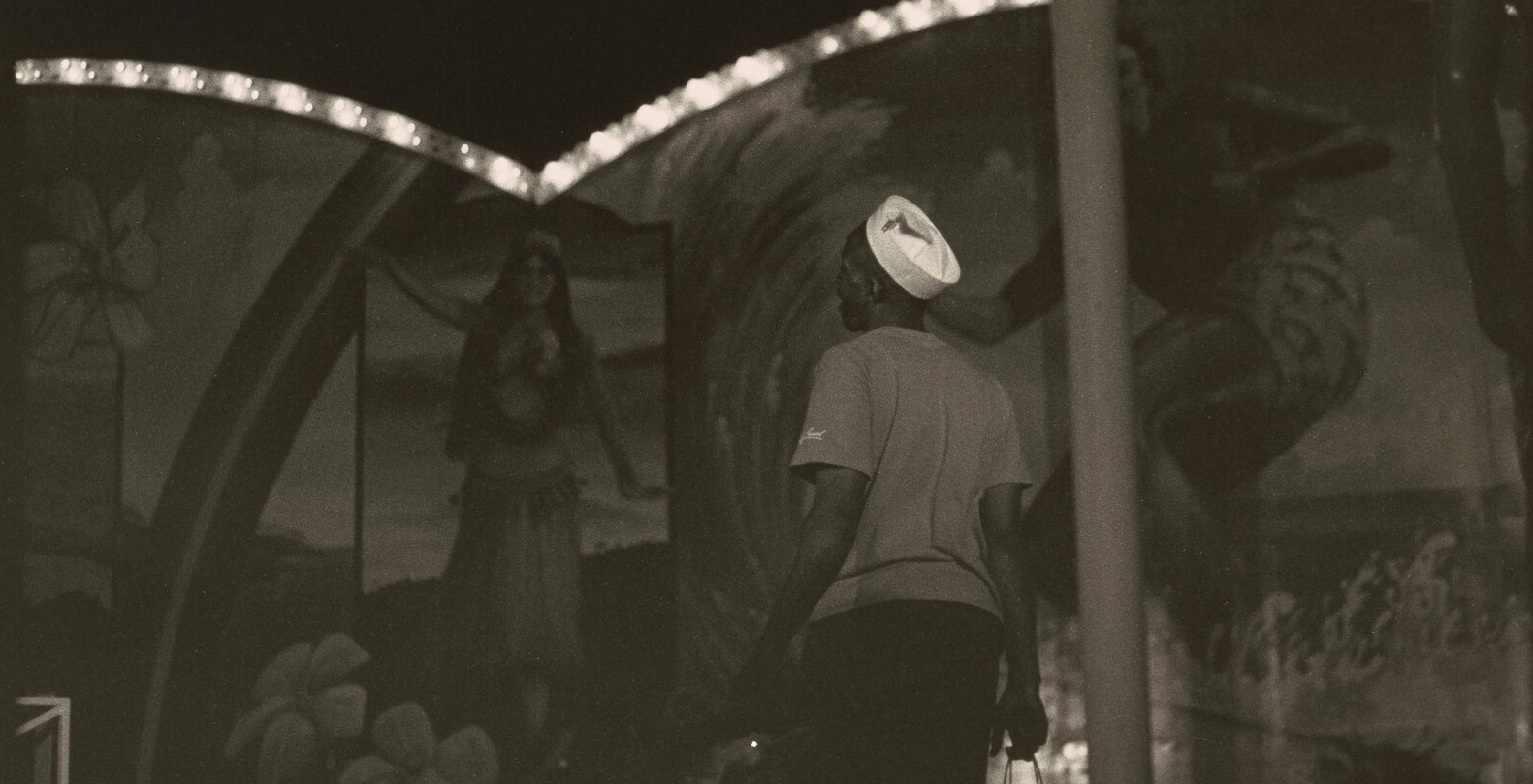 Photograph of a man walking away from the camera with circus lights in the background