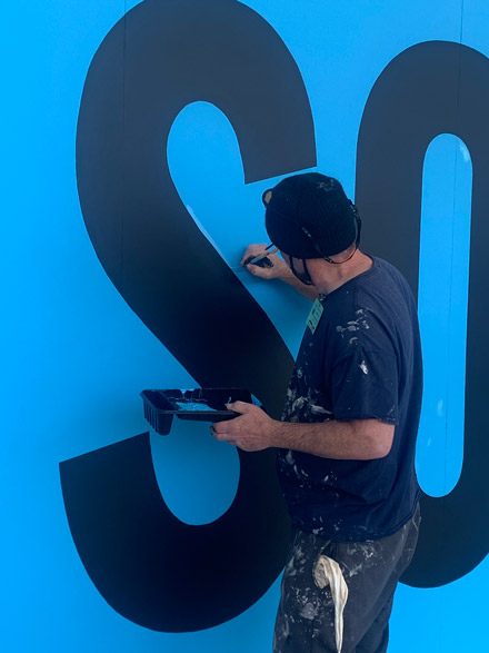 Man touching up the outline of the blue background on the painting SORRY by Kay Rosen
