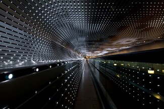 We look through a tunnel lined with dozens of strings of bright white lights nestled in silver-gray slats that zoom away from us in this horizontal photograph. At the far end of the tunnel, at the center of the photograph, the alternating lines of lights and slats almost come together around a narrow, capsule-shaped area of golden yellow light. The flat ceiling curves down to meet the wall to our left. The curve continues to make a C-shape before extending straight down the remaining height of the wall, turning the profile of the open space into a backward-facing P. Some of the lights are bright, some fading, and some are off to create a loose pattern of dark squares floating in a field of light. The lights seem brighter at the far end of the tunnel. The overall impact of the view is a starburst radiating in diagonal lines, coming from a point at the center of the photograph.