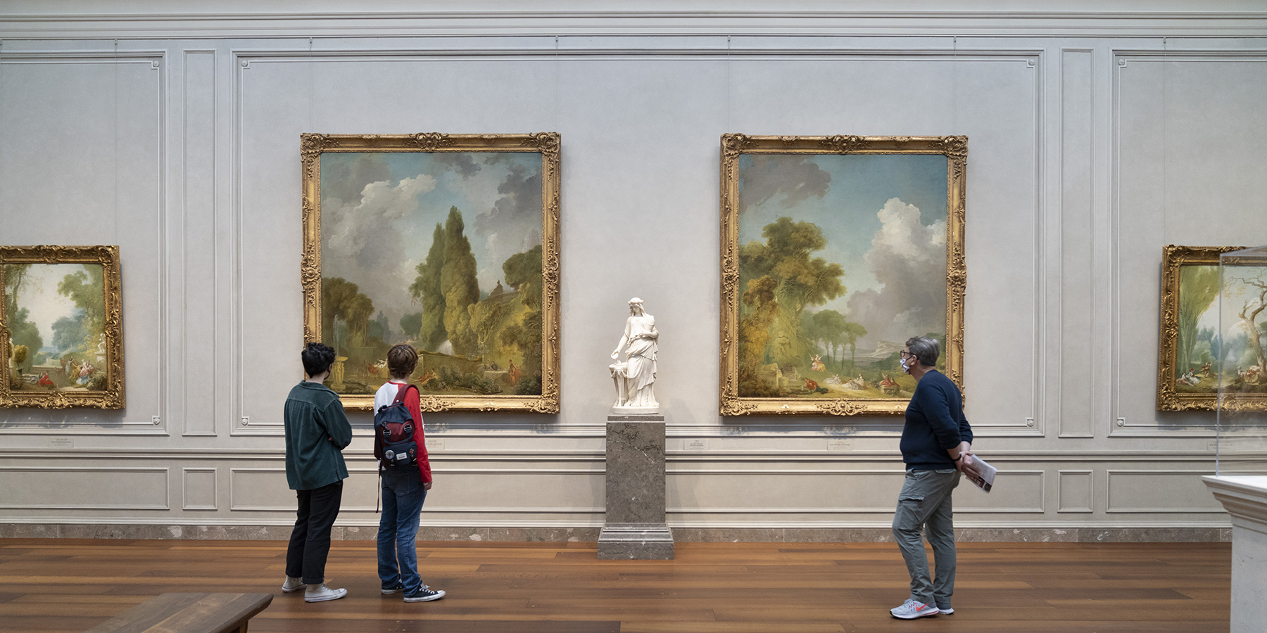 A woman with a facemask on and arms crossed is looking at a sculpture that displayed on a pedestal along with other sculptures that are also on pedestals. There is a man in the background taking a picture of another sculpture on his phone.