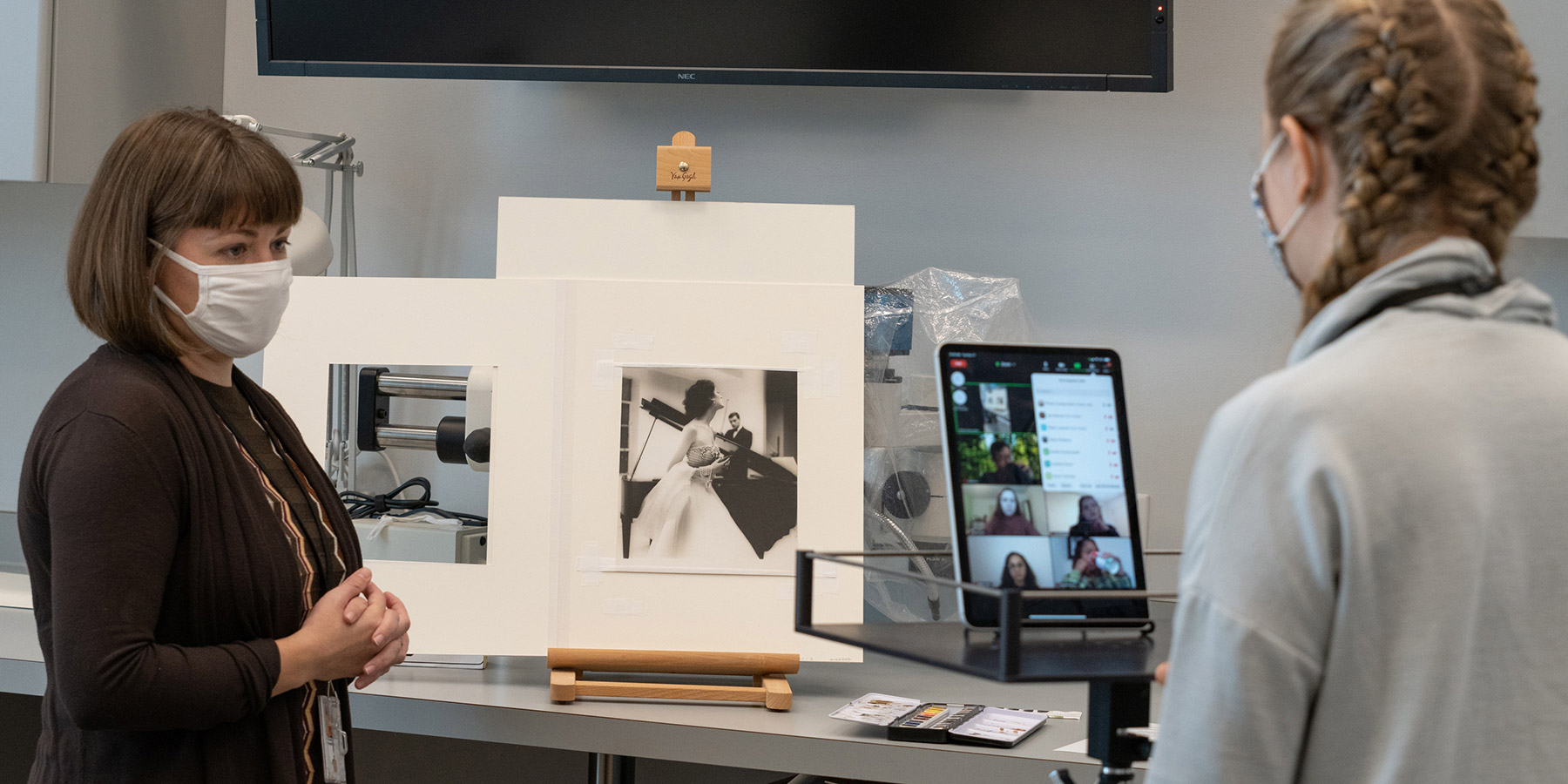 A masked person standing in front of a photograph on an easel is giving a virtual presentation via iPad while another person with a facemask looks at tablet