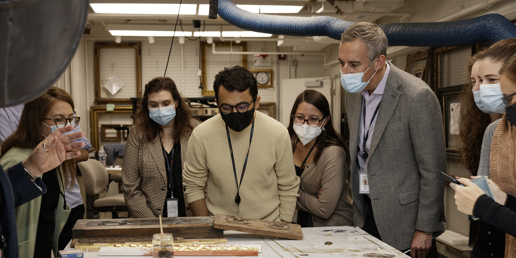A masked person standing in front of a photograph on an easel is giving a virtual presentation via iPad while another person with a facemask looks at tablet