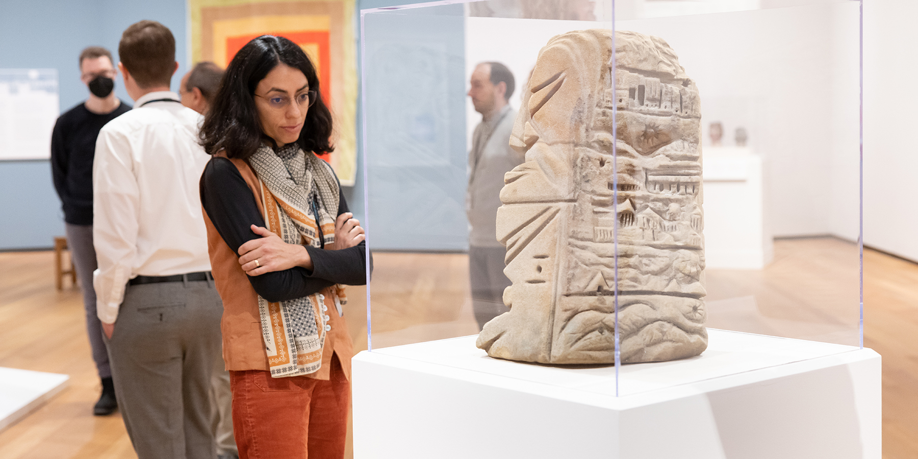 A masked person standing in front of a photograph on an easel is giving a virtual presentation via iPad while another person with a facemask looks at tablet