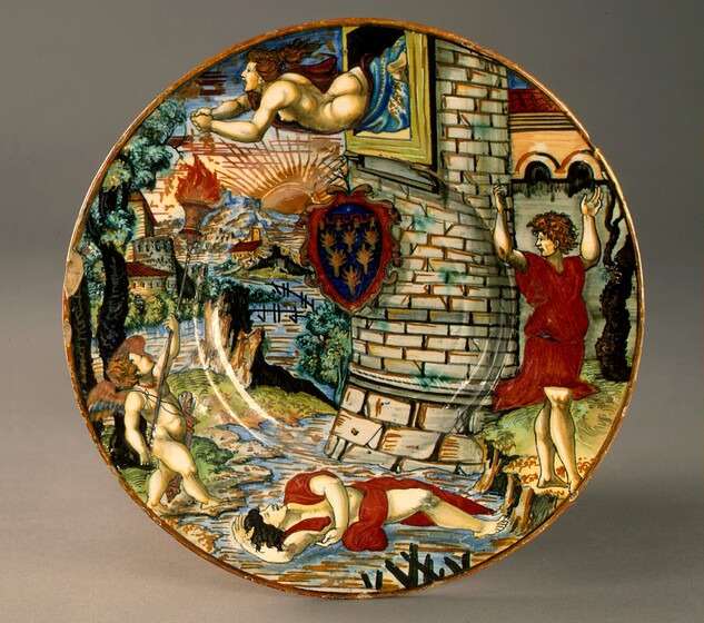 This circular plate is painted with an almost cartoon-like scene showing four people with pale skin around a stream, near the exterior of a tower. At the top of the plate, a nude woman is shown from the thighs up as she falls horizontally from an open window at the top of the tower. Her back, buttocks, and a single breast are exposed to us while a deep red and blue fabric wraps over her far shoulder and around her thighs. Her hands clasp as she reaches her arms forward, and her copper-red hair flies behind her. Her mouth is wide open. Directly below her, at the bottom center of the plate, a man partially wrapped in ruby-red drapery lies on his back in the stream. One arm is flung over his head and the other hangs across his body. He has black hair, and his eyes are closed. On the right side of the plate, a second man stands on the bank of the stream with his body facing us, as he turns his face to our left in profile. He has curly, brown hair, and the eyebrow we can see is furrowed. He wears a knee-length red tunic, and he raises both arms high overhead. Finally, the fourth winged, child-like person stands on the opposite bank, to our left. He is nude except for a quiver of arrows slung across his chest. His babyish body is chubby with small wings between his shoulder blades. He faces the center of the scene and looks up at the falling woman while raising a long torch toward her. The tower is made of parchment-white stones outlined with mustard yellow and black. A shield or coat of arms hangs or hovers just above the center of the plate. It has a rounded top and pointed bottom, and has six gold, tree-like forms against a navy-blue background, all within a scrolling, red frame. The stream winds into the distance between buildings along the banks. Cobalt-blue and sepia-brown mountains line the horizon, and a light brown sun with linear rays sets behind them. The plate is edged with a thin band of golden brown, and there are several chips around the rim. The plate is shown in front of a light gray background and it casts a light shadow to our right.