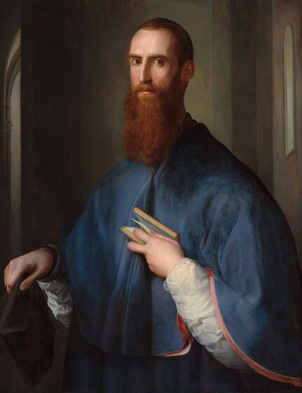 A slender, light-skinned man with short brown hair and a long chestnut beard is shown from the hips up, his chest and sloping shoulders covered by a soft cape of muted, cobalt blue, and he turns an intense brown-eyed gaze toward us in this vertical portrait painting. He nearly fills the composition, with his right shoulder, on our left, drawn back slightly as he looks straight at us from the corners of his eyes under dark, low brows. His head, torso, aquiline nose and hands are elongated. He holds a black hat in his right hand, on our left, close to his body. His left hand emerges from a radiant white sleeve painted with swirling brushstrokes, as he presses a small book to his chest, holding it open with one page caught between thumb and index finger. The lower edge of his cape drapes over his raised left forearm, revealing a pink lining that forms a curves in a shallow C toward the lower right corner. He stands with his back to a greyish-green wall, shadowed on his right, our left. The wall behind his opposite shoulder is lit by the same light that illuminates his face from our left. Slivers of other spaces, possibly rooms, are suggested by tall, narrow views of windows and arched openings along the sides of the painting.