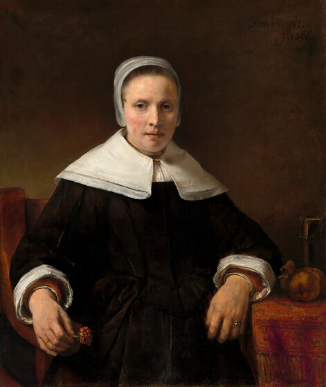 Shown from the lap up against a dark background, a pale-skinned woman wearing a black dress looks out at us in this vertical portrait. Soft light coming from our left highlights her oval face, the peach blush on her cheeks, and her crisp white collar, cap, and cuffs. Her brown hair is pulled back under the cap that flares slightly over her ears and the wide, lace-trimmed collar covers her shoulders. She has dark eyes, a straight nose, and her pink lips are slightly parted. Her left arm, on our right, rests on a table covered with a rose-red patterned carpet next to two pieces of fruit, perhaps apples, and a book fastened with metal clasps. She rests her other arm on the arm of the wooden chair and holds a deep pink carnation the same color as the carpet. She wears a ring on the third finger of each hand. The artist signed the work with black paint against the dark background near the upper right corner: “Rembrandt. f.1656.”